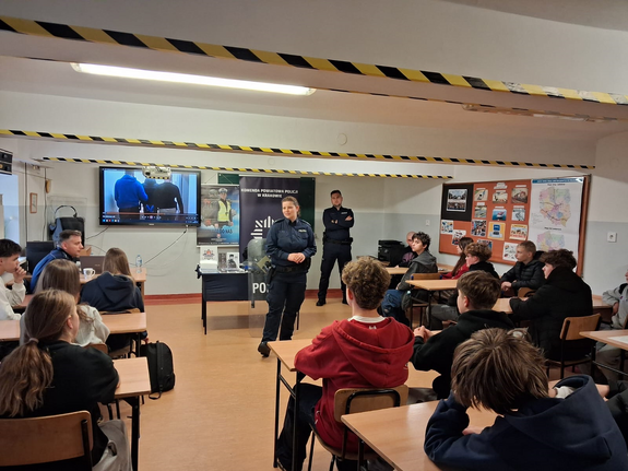 policjanci w mundurach w sali podczas prelekcji przed nimi siedzą w ławkach uczniowie