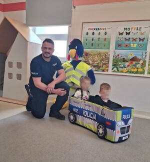 policjant w mundurze obok maskotka policyjna i dwójka dzieci w autku