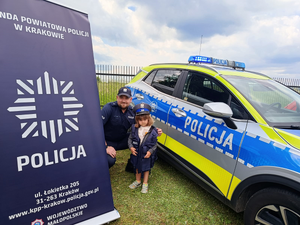 policjant w mundurze z dziewczynką w policyjnej czapce obok radiowóz i baner komendy