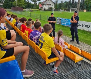 policjanci w mundurach przed nimi siedzą uczniowie na trybunach na boisku sportowym
