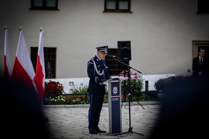 policjant w mundurze stojący przy mównicy  przemawiający do zgromadzonych funkcjonariuszy na dziedzińcu