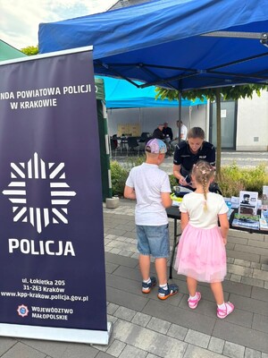 baner KPP Kraków dzieci przy stoliku i policjantka w mundurze rozmawiająca z dziećmi i prezentująca im materiały edukacyjne