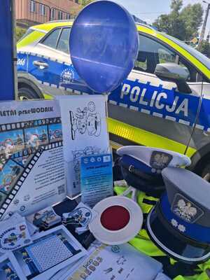 materiały edukacyjne w formie ulotek ,granatowy balon z logo policji ,czapki policjantów ruchu drogowego z białymi otokami , wszystko to rozłożone na stoliku, w tle również radiowóz oznakowany