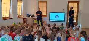 policjantka i policjant w mundurach w sali przedszkolnej przed nimi siedzą dzieci