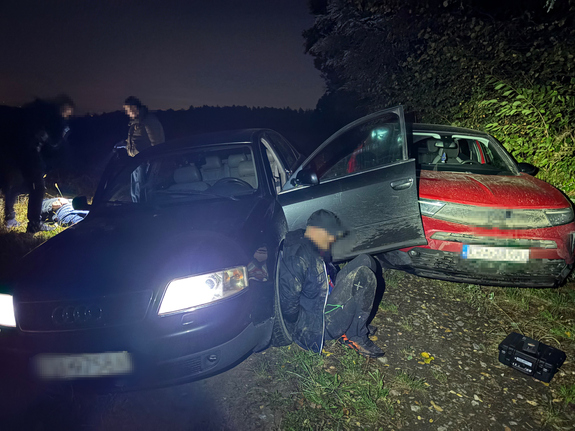 dwa samochody na polnej drodze przy jednym aucie na ziemi siedzi zatrzymany skuty kajdankami mężczyzna
