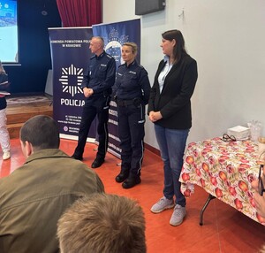 policjant i policjantka w mundurach obok nich stoi kobieta po cywilnemu w sali podczas prelekcji przed nimi siedzą seniorzy