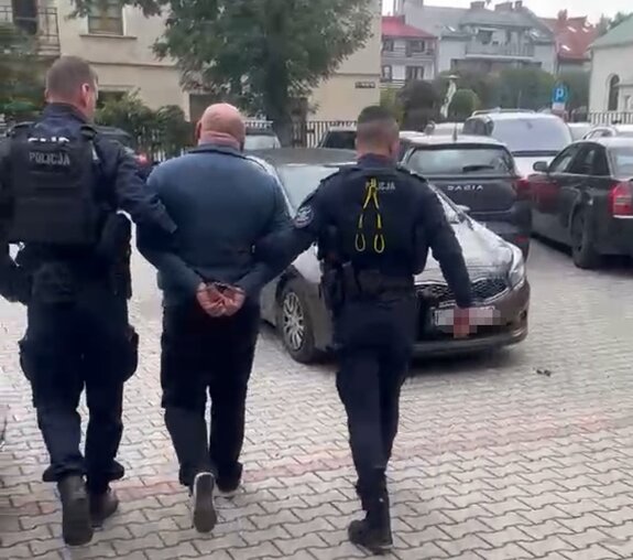 policjanci w  mundurach prowadzą zatrzymanego do radiowozu na parkingu komisariatu