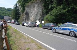 radiowóz i ciąg aut zaparkowanych przed nim,w tym karetka, po drugiej stronie za ogrodzeniem stojący ludzie