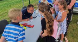 policjant przy stanowisku policyjnym na pikniku, wokół niego dzieci z którymi rozmawia, w tle radiowóz