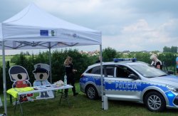 radiowóz i idąca policjantka ustawiona tyłem, w tle ludzie i stoisko z policyjnymi gadżetami