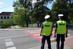 dwóch umundurowanych policjantów z ruchu drogowego stających przed przejściem dla pieszych, w tle szkoła