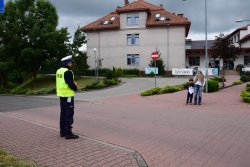 umundurowany policjant stojący przy drodze, po drugiej stronie jezdni kobieta z dzieckiem, w tle szkoła