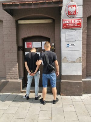 stojący tyłem policjant doprowadzający zatrzymanego z kajdankami założonymi na ręce trzymane z tyłu do aresztu śledczego