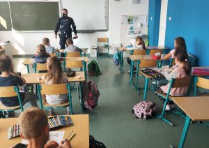 policjant stojący na środku sali przeprowadzający w klasie prelekcje dla pierwszoklasistów, siedzących w ławkach