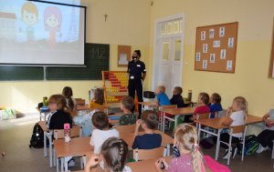 dzieci siedzące w ławkach, widoczne tyłem, oglądające podczas prelekcji film edukacyjny, przy ekranie stojąca policjantka