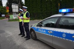 dwóch umundurowanych policjantów stojących przy radiowozie na poboczu, jeden z nich trzyma gadźety i elementy odblaskowe promujące bezpieczeństwo