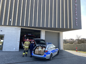 strażak z OSP z policjantką, obok radiowóz z otwartym bagażnikiem wypełniony torbami z  produktami żywnościowymi, w tle budynek OSP Wielka Wieś