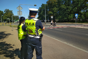 dwoje policjantów stojących tyłem do zdjęcia a przodem do drogi, w tle kobieta z wózkiem przechodząca przez drogę