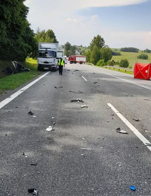 miejsce wypadku drogowego, policjant ruchu drogowego stojacy przy pojeżdzie cięzarowym, w rowie po lewej leżący zmiażdżony pojazd, po drugiej stronie namiot za którymi leżą zwłoki, w tle pojazd strażacki