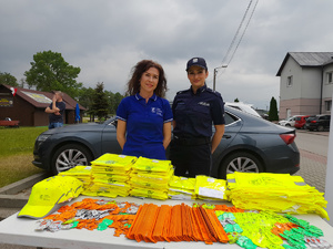 przedstawicielka starostwa powiatowego z policjantką stojące przy stoisku z elementami odblaskowymi, obok baner starostwa , za nimi w tle pojazd..