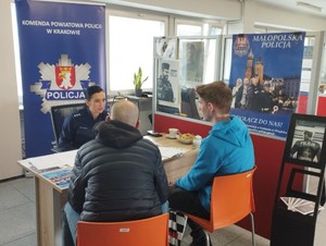 policjantka opowiadająca o służbie siedzącym po przeciwnej stronie biurka dwóm mężczyznom, za policjantką baner KPP Kraków, widoczne też ulotki na stojaku