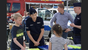 dwoje umundurowanych policjantów  stojących przy stoisku policyjnym, na którym leżą elementy odblaskowe, wokół dwoje dzieci z którymi policjantka rozmawia, w tle wóz strażacki