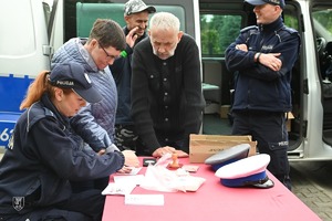 policjantka przy stoisku, odbijająca linie papilane na papier uczestniczce imprezy, za nimi policjant rozmawiajacy z mężczyzną, w tle radiowóz