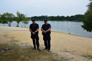 dwóch policjantów stojących przodem na plaży, przed nimi zalew w którym pływają kajaki