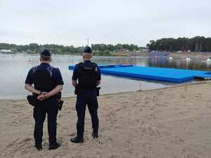 dwóch policjantów stojących tyłem do zdjęcia, przed nimi zalew na którym odbywają się zawody, w tle po prawej trybuna na plaży