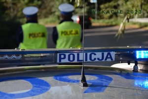 dwóch policjantów ruchu drogowego stojących przed radiowozem i obserwujących drogę I NAPIS WAKACJE 2023