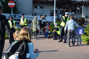 polciajnci i strażniczka gminna rozdają odblaski przy przejściu dla pieszych, w tle szkoła i ludzie oraz uczniowie