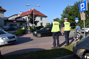 policjanci stojący tyłem, przed nimi przejście dla poieszych, w tle szkoła i przechodnie