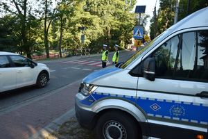 zaparkowany bok radiowozu przy drodze, w tle policjanci stojący przy przejściu dla pieszych i jadące samochody