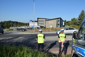 dwóch policjantów ruchu drogowego stojący tyłem, obok nich radiowóz, przed nimi w tle szkoła