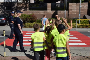 dwoje policjantów przeprowadza dzieci z podniesionymi rękami, ubrane w kamizelki odblaskowe przez jezdnię