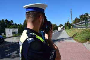 policjant od ramion w górę, stojacy tyłem do zdjęcia, mierzący miernikiem prędkości w jadące popjazdy