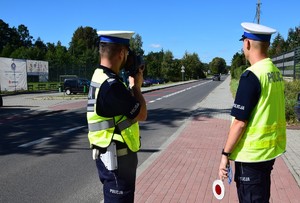 dwóch policjantów ruchu drogowego jeden trzyma miernik prędkości i mierzy w jadący pojazd, drugi trzyma w ręku lizaka do zatrzymywania pojazdów