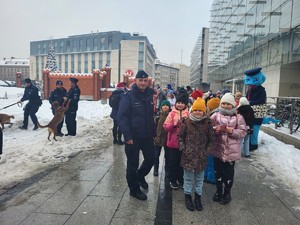 1. dzieci stojace w parach, obok nich zastępca komendanta KPP w Krakowie, w tle po lewej policjanci z psem policyjnym