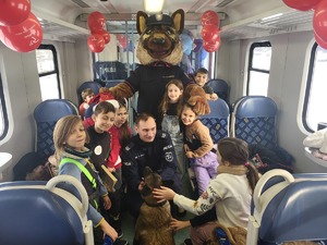 6 maskotka strazy ochrony kolei, przed nim policyjny pies z przewodnikiem, którego głaskają dzieci w pociągu
