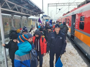 8 policjantka stojaca na dworcu przy pociągu, obok niej i za nią stojące dzieci, w tle maskotki policyjne