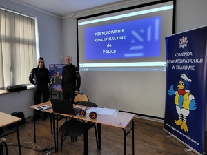 dwoje umundurowanych fucnkjonariuszy stojacych przy wyświetlonej prezentacji, miedzy nimi plakat dot. rekrutacji do Policji, po prawej stronie baner komendy.