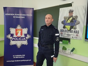 policjant umundurowany stojący w sali, obok niego baner KPP Kraków, za nim wiszący na tablicy plakat promujący zawód policjanta