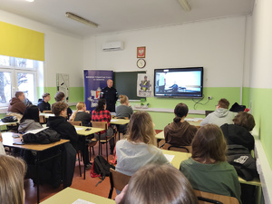 siedzący w ławkach uczniowie, przed nimi policjant prowadzący prelekcje, za nim wyświetlona prezentacja